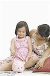 Girl putting a coin into a piggy bank with her mother sitting behind her and smiling