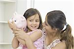 Side profile of a girl holding a piggy bank with her mother looking at her and smiling