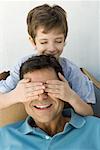 Boy with hands over his father's eyes, both smiling, boy's eyes closed