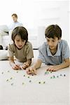 Boys lying on the floor playing with marbles, father sitting on sofa with laptop in background