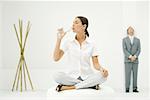 Professional woman sitting on ottoman, drinking from water bottle, businessman and bamboo in background