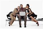 Businessman using a laptop and two businesswomen leaning on his shoulders