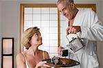 Senior man pouring tea from a tea kettle into the cup with a mature woman holding a tray