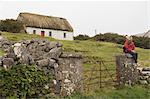 Frau sitzt auf Tor vor traditionellen Reetdachhaus in Inishmor, Aran Islands, County Galway, Irland