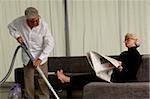 Senior man vacuuming, senior woman sitting on sofa while reading a newspaper