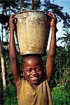 Kopf und Schultern Porträt eines jungen afrikanischen Kindes trägt einen Eimer Wasser auf dem Kopf, lächelnd und Blick in die Kamera, Liberia, Westafrika, Afrika