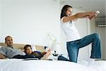 Girl on bed playing video game with wireless controller, parents watching in background