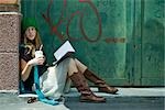 Young woman sitting on ground with book and drink, looking away