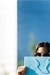 Young woman wearing sunglasses, holding shopping bag in front of face