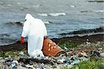 Person in protective suit carrying barrel of hazardous waste on polluted shore