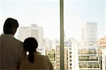 Couple, woman's head on man's shoulder, together at window looking at view of city