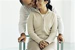 Woman sitting in chair, husband standing behind leaning down whispering in her ear, front view