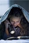 Girl laying in bed under blanket with flashlight reading