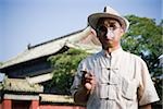 Man with sunglasses and straw hat smoking cigarette outdoors with pagoda in background