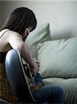 Woman sitting on sofa with laptop and headphones playing guitar