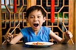 Boy sitting at table with empty plate holding chopsticks scowling