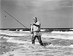 HOMME PLUS ÂGÉ DES ANNÉES 1950 AU SURF EN CUISSARDES TENANT UN POISSON EN UNE MAIN CANNE À PÊCHE DANS D'AUTRES