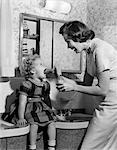 1950s MOTHER ADMINISTERING MEDICINE WITH SPOON TO YOUNG DAUGHTER WITH MOUTH WIDE OPEN SITTING ON BATHROOM SINK