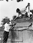 1930s PAIR OF BOYS BUILDING SHACK ONE NAILING BOARD TO SIDE ONE ON ROOF WITH SAW