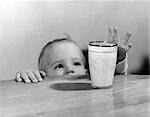 1950s TODDLER REACHING UP TO GRAB GLASS OF MILK ON TOP OF TABLE