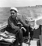1950s YOUNG TEEN BOY STEERING BOAT WITH HIS HAND ON OUTBOARD MOTOR AND HIS FOOT ON TILLER WEARING WHITE SAILOR HAT