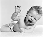 1950s BABY LYING ON STOMACH LAUGHING WITH SQUINTED EYES & HAND IN AIR