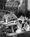 1960s SUBURBAN FAMILY OF FOUR AT PICNIC TABLE IN BACKYARD EATING