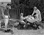 1960s MAN ON PATIO GRILLING STEAK WITH 2 DAUGHTERS SEATED AT PICNIC TABLE & WIFE STANDING SERVING FOOD