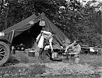 1920s 1930s COUPLE MAN WOMAN ON FISHING CAMPING TRIP VACATION AT CAMPSITE MAN COOKING OVER FIRE