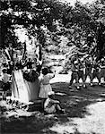 1950s A BOY AND A GIRL STAND BY A TOMBSTONE WAVING TO PASSING COLOR GUARD OF SOLDIERS IN A MEMORIAL DAY PARADE