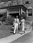 1930s COUPLE WALKING OUT OF HOUSE DOWN SIDEWALK CARRYING PICNIC BASKETS & THERMOS JUG