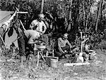 1940s MEN AT CAMPSITE ONE WASHING HIS FACE AT TRIPOD STAND THE OTHER TENDING CAMPFIRE CANADA LAKE OF THE WOODS ONTARIO