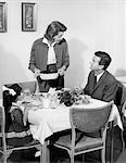 1960s FAMILY OF THREE AT DINING ROOM TABLE AT DINNER TALKING WITH WIFE STANDING PUTTING CASSEROLE DISH ON TABLE