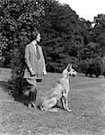 DES ANNÉES 1940, FEMME PORTANT LE COSTUME DE TWEED RIDING BOTTES DEBOUT AVEC PROFIL DE DOGUE ALLEMAND EN PLEIN AIR