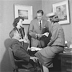 1950s TWO MEN AND TWO WOMEN DISCUSSING FABRIC SAMPLE AT DESK