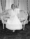 1970s WOMAN WEARING WHITE FUR TRIMMED DRESSING GOWN SITTING ON STRIPED LOVE SEAT INDOOR LOOKING AT CAMERA