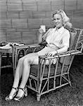 1940s BLOND WOMAN IN RATTAN CHAIR HOLDING UP GLASS OF MILK SMILING WEAR SHORTS WHITE SHIRT AND STRAP OPEN TOED SHOES