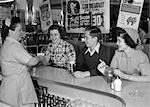 ANNÉES 1950 FEMME SOURIANTE SERVEUSE QUI DESSERT LES DEUX JEUNES FILLES ET UN GARÇON AU COMPTOIR DE LA SALLE À MANGER