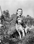 ANNÉES 1940 FEMME TENANT UN FUSIL DANS UNE MAIN CARESSER CHIEN AVEC L'AUTRE À GENOUX DANS L'HERBE AVEC COSTUME DE TWEED