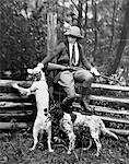 FEMME DES ANNÉES 1920 EST ASSIS SUR LA CLÔTURE EN BOIS TENANT LE FUSIL DE CHASSE SUR UN GENOU CARESSER UN DES DEUX CHIENS DE CHASSE CHAPEAU FASHION