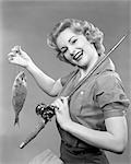 1950s SMILING WOMAN WITH A FISHING ROD OVER HER SHOULDER HOLDING UP A FISH