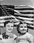 1940s PATRIOTIC COMPOSITE OF SMILING BOY AND GIRL OVER BACKGROUND OF THE AMERICAN FLAG BLOWING IN THE WIND