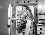 1950s WOMAN WEARING GINGHAM DRESS AND APRON OPENING REFRIGERATOR DOOR