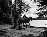 ANNÉES 1930 HOMME & FEMME CHEVAL CHEVAUX À TRAVERS UNE ZONE BOISÉE LE LONG DE LA RIVE DU LAC