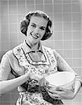 1950s SMILING WOMAN WEARING APRON IN KITCHEN HOLDING MIXING BOWL AND SPOON LOOKING AT CAMERA