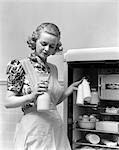 1930s WOMAN WEARING A WHITE APRON TAKING MILK OUT OF THE REFRIGERATOR