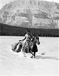 1930s COWBOY WITH BATWING CHAPS ON A BAY HORSE CROSSING A RIVER LEADING A PAINT PACK HORSE WITH MOUNTAINS IN BACKGROUND