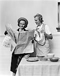 1940s 2 WOMEN IN KITCHEN ONE DRYING DISHES WITH TOWEL OTHER IN HAT COAT HOLDING READING NEWSPAPER