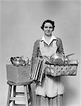 1930s 1940s WOMAN HOUSEWIFE FACING CAMERA HOLDING BASKET FILLED WITH POTS PANS COOKWARE CHORE APRON