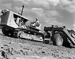 1950s ANGLED WORM'S-EYE VIEW OF MAN PULLING LEVERS ON BULLDOZER HAULING SCRAPER